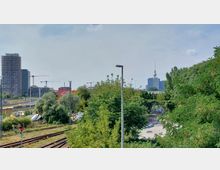 Städtische Landschaft mit Blick auf Bahngleise im Vordergrund, dichtem Grün und Bäumen in der Mitte sowie modernen Hochhäusern und Baukränen im Hintergrund; rechts ist der Berliner Fernsehturm sichtbar.