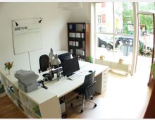 Helles Büro mit einer großen Fensterfront, die auf eine Straße blickt. Im Raum stehen weiße Schreibtische mit Computerbildschirmen, Büromaterialien und einer Schreibtischlampe; links ein Regal mit Drucker und Aktenordnern, dahinter ein schwarzes Regal.