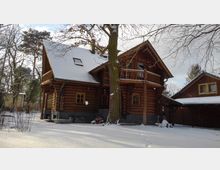 Holzhaus mit Schnee bedeckt, umgeben von einer winterlichen Landschaft mit kahlen Bäumen. Das zweistöckige Gebäude hat ein Satteldach, einen kleinen Balkon und Holzfenster; daneben ist ein weiteres Holzgebäude sichtbar.