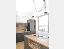 Moderne Küche mit dunklen grauen Schränken und Holzarbeitsflächen. In der Mitte befindet sich eine Kücheninsel mit integriertem Kochfeld und abgesetztem Regal für Weingläser; große Fenster lassen viel Tageslicht herein.