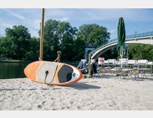 Sandstrand mit einem orange-weißen Stand-Up-Paddle-Board im Vordergrund, daneben mehrere gestapelte Klappstühle und geschlossene Sonnenschirme. Im Hintergrund befinden sich grüne Bäume, ein Fluss sowie eine geschwungene Brücke mit einem kleinen Turm.