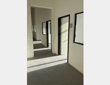 Heller Innenflur in einem modernen Gewerbe- oder Bürogebäude mit weißen Wänden und sichtbaren Ziegelpfeilern, grauem Boden sowie Türen und Fenstern mit schwarzen Rahmen. Sonnenlicht fällt durch die Fenster und wirft lange Schatten auf den Boden.