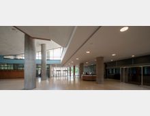 Großzügige Empfangshalle mit glatten Bodenfliesen, runden Betonpfeilern und einer abgehängten Decke mit eingelassenen Lichtern. An einer Seite befindet sich ein runder Empfangstresen, und die Wände sind teilweise mit Glasfronten und Holzelementen verkleidet, während große Fenster Tageslicht hereinlassen.