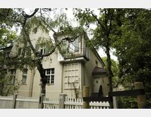 Zweistöckiges Wohnhaus mit hellem Putz und Satteldach, umgeben von dichtem Baumbestand. Vor dem Haus befindet sich ein weißer Lattenzaun und eine kleine Treppe führt zur Eingangstür.