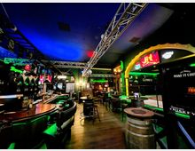 Innenansicht einer Bar im Pub-Stil mit geschwungener Theke, hohen Barhockern und mehreren Stehtischen auf Holzboden. Grüne und blaue Akzentbeleuchtung, Metall-Traversen an der Decke sowie Werbeschilder und Dekorelemente prägen den Raum.