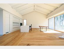 Minimalistischer Wohnraum mit heller, offener Küche, weißen Einbauschränken und einer zentralen Kücheninsel. Der Raum hat eine Holzdecke mit sichtbaren Balken, einen Holzboden und eine große Fensterfront mit Blick auf eine Außenlandschaft.