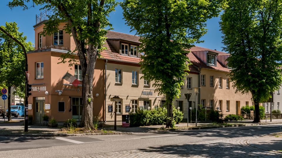 Straßenecke mit einem zweigeschossigen, beigefarbenen Gebäude mit rotem Ziegeldach und Gauben, umgeben von hohen Straßenbäumen. Im Vordergrund sind Kreuzung, Gehwege, Verkehrszeichen und Ampelmasten zu sehen, rechts schließt sich eine Reihe ähnlicher Wohn- und Geschäftshäuser an.