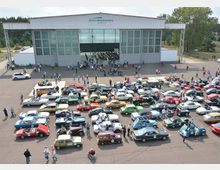 Eine Luftaufnahme eines Parkplatzes mit zahlreichen klassischen Autos in verschiedenen Farben und Modellen, die von Personen umgeben sind. Im Hintergrund befindet sich ein großes Gebäude mit der Aufschrift „Airport Neuhardenberg“, das wie ein Hangar gestaltet ist, umgeben von Bäumen und einer offenen, ebenen Landschaft.