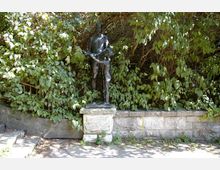 Bronzestatue eines Mannes und eines Kindes auf einem steinernen Sockel, umgeben von dichtem grünen Blattwerk und einer niedrigen Steinmauer im Hintergrund. Links im Bild sind Stufen sichtbar, die zur Statue führen.