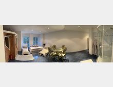 Ein geräumiges Zimmer mit einem dunklen Fliesenboden und heller Wandgestaltung. Links steht eine Holzsauna, daneben ein Bereich mit zwei Betten und Sesseln vor bodentiefen Fenstern. Rechts im Bild befindet sich eine Glasdusche und daneben Bademäntel an Haken sowie ein runder Glastisch mit vier Stühlen in der Mitte des Raumes.