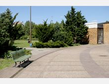 Ein gepflasterter Weg in einem Park, flankiert von grünen Büschen und Bäumen. Auf der linken Seite steht eine grüne Holzbank, während rechts ein Gebäude mit einer Fassade aus gelben Ziegelsteinen und großen Fenstern sichtbar ist.