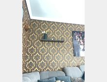 Wohnzimmer mit einer gold-schwarz gemusterten Tapete im Barockstil, einem grauen Ecksofa mit Kissen, einem Wandregal mit Dekoration wie einer Vase und kleinen Objekten sowie einem großen gerahmten Blumenbild an der Wand.