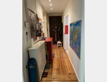 Langer, schmaler Hausflur mit Holzfußboden und weißen Wänden, dekoriert mit einer Weltkarte und einem gerahmten Poster. Der Flur enthält eine weiße Kommode, ein rotes Sideboard, diverse Schuhe und ein an der Wand aufgehängtes Skateboard; am Ende sind dunkle Holztüren zu sehen.