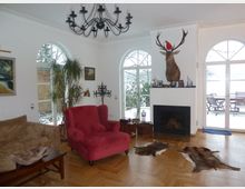 Wohnzimmer mit Parkettboden, einem Kamin in der Mitte der Wand, auf dem ein Hirschkopf mit Weihnachtsmütze montiert ist. Rundbogenfenster und Glastüren lassen viel Tageslicht herein, Möbel umfassen ein rotes Polstersessel, ein braunes Sofa und Tierfelle auf dem Boden.