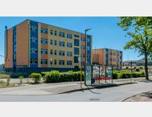 Gelbes Schulgebäude mit blauen Fassadenelementen, vier Stockwerken und zahlreichen rechteckigen Fenstern. Im Vordergrund steht eine überdachte, verglaste Bushaltestelle mit Graffiti sowie umliegendem Grün und Gehwegen.