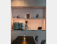 Moderne Kaffee-Ecke mit einer silbernen Espressomaschine und einer Kaffeemühle auf einer hellen Arbeitsplatte. Darüber befinden sich Regale mit dekorativen Gegenständen, Flaschen und einer Nespresso-Kapselaufbewahrung; im Vordergrund stehen schwarze Tische mit kleinen Kerzen.