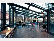 Modernes Wohnzimmer mit Glaswänden und gläsernem Dach, ausgestattet mit schwarzen Ledersesseln, einem roten Sofa, einem gläsernen Esstisch und Bücherregalen auf der linken Seite. Der Raum bietet einen Blick auf umliegende Häuser und Bäume.