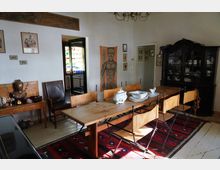 Esszimmer mit einem langen Holztisch, umgeben von Stühlen mit Lederbespannung, auf einem roten Teppich. An der Wand hängen Bilder und ein großes Gemälde, daneben ein dunkler Holzschrank mit Geschirr sowie ein Sideboard mit einer Buddha-Statue. Im Hintergrund führt eine Tür in einen Raum mit farbigen Glasflaschen.