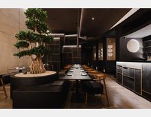 Ein modernes Restaurant mit dunkler, minimalistischer Inneneinrichtung, zentralem Bonsai-Baum in einem großen Pflanzgefäß und langen, gedeckten Tischen mit Holzmöbeln. Die Wände sind dunkel mit goldenen Akzenten, ergänzt durch dezente Beleuchtung und kreisförmige dekorative Elemente.