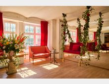 Heller Salonraum mit großen Sprossenfenstern und roten Vorhängen, Holzboden sowie Sitzmöbeln in Rot. In der Mitte hängt eine dekorative Schaukelbank, umrankt von grünen Ranken mit roten Rosen; daneben stehen ein kleiner Beistelltisch und ein großer Blumenstrauß auf einer Steinsäule.