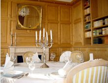 Elegantes Speisezimmer mit holzgetäfelten Wänden, einem Kamin mit Spiegel darüber, und Bücherregalen an der Seite. Ein großer, rund gedeckter Tisch ist mit Kerzenhalter, gefalteten Servietten und Weingläsern geschmückt.