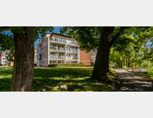 Mehrstöckiges Wohngebäude mit Balkonen, umgeben von einer Rasenfläche und großen, schattenspendenden Bäumen. Rechts führt ein gepflasterter Weg entlang einer Parkbank durch die grüne Umgebung.