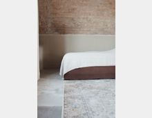Minimalistisch eingerichtetes Schlafzimmer mit einer unverputzten Backsteinwand, einem niedrigem Bettgestell aus Holz mit weißer Bettwäsche und einem großen, gemusterten Teppich auf einem Betonboden.