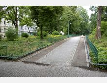 Gepflasterter Weg in einem Park mit grünen Metallzäunen auf beiden Seiten, umgeben von dichter Vegetation und Bäumen. Im Hintergrund sind Laternen und ein großer Stein zu sehen; links am Rand stehen Gebäude.
