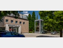 Außenansicht einer Stadthalle mit moderner Fassade aus beigen Paneelen, großen Glas-Eingangstüren und dem Schriftzug „Stadthalle“. Rechts davon steht eine Glasvitrine mit einer Statue, umgeben von Bäumen und geparkten Autos.