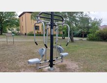 Outdoor-Fitnessgerät auf einer Wiese in einem Parkbereich mit vereinzelten Bäumen und Büschen, im Hintergrund ist ein gelbes Wohngebäude sichtbar. Ein schmaler Weg führt durch das Gelände.