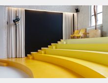 Halbkreisförmiger, gelber Treppenbereich in einem modernen Innenraum, mit hellen Vorhängen, einer schwarzen Wand im Hintergrund und zwei gelben Klappstühlen auf der oberen Ebene. Rechts steht ein Fenster mit einem Heizkörper und einer hölzernen Pflanzenhalterung mit einer Palme.