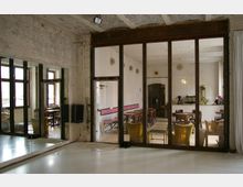 Innenansicht eines Gebäudes mit rohen, unverputzten Wänden und einer großen Glaswand, die einen rustikalen Raum mit Holzmöbeln und einer kleinen Bar abtrennt. Der Vordergrund zeigt einen Bereich mit Spiegelwand und poliertem Boden, möglicherweise für Tanz- oder Bewegungszwecke.