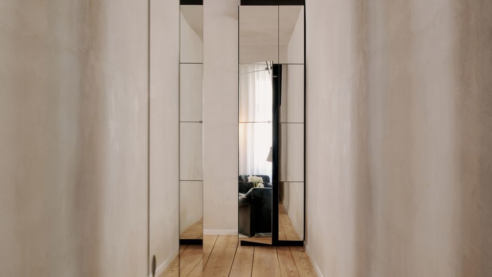 Schmaler, minimalistischer Flur mit hell verputzten Wänden, breiten Holzdielen und dunkler Decke. Am Ende stehen hohe Spiegeltüren, die den angrenzenden Wohnbereich und ein Fensterlicht reflektieren.