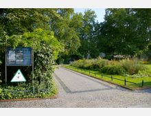 Gepflasterter Weg in einem kleinen Park mit Blumenbeeten, umgeben von hohen Bäumen. Links steht eine Infotafel mit der Aufschrift „Savigny-Platz“, daneben ein Hinweisschild für geschützte Pflanzen.
