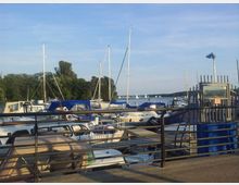 Eine Marina mit mehreren festgemachten Segel- und Motorbooten, einige mit blauen Abdeckplanen. Im Hintergrund ist ein See mit kleinen Segelbooten und bewaldeter Uferlandschaft unter einem sonnigen Himmel zu sehen.