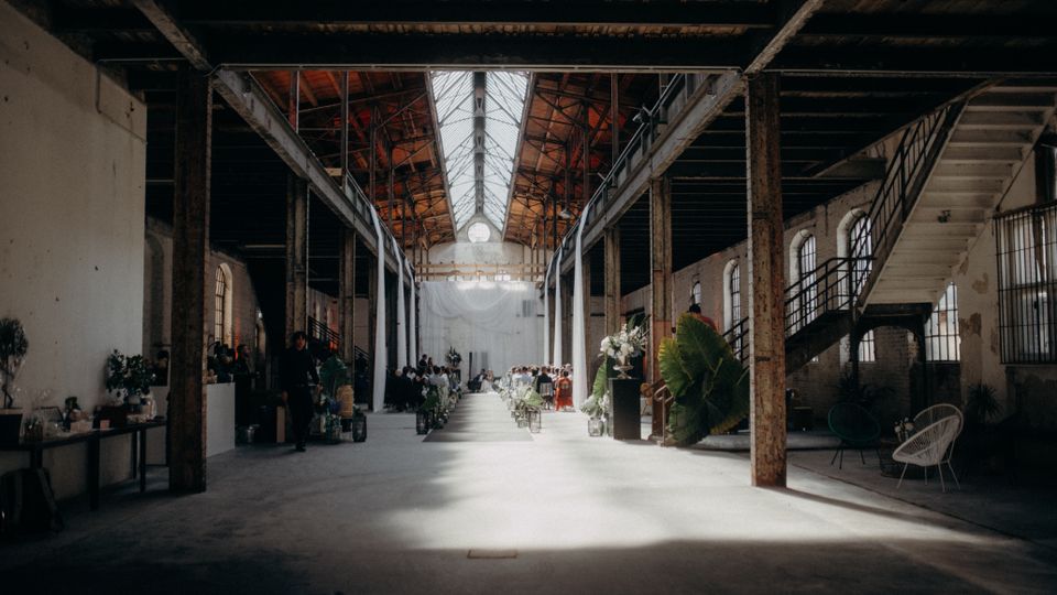 Großer, industrieller Innenraum einer alten Fabrikhalle mit sichtbaren Metallträgern und Holzdecken. In der Mitte führt ein geschmückter Weg zu einer Bühne, während auf beiden Seiten Sitzplätze und Dekorationen mit Pflanzen und Blumen zu sehen sind.