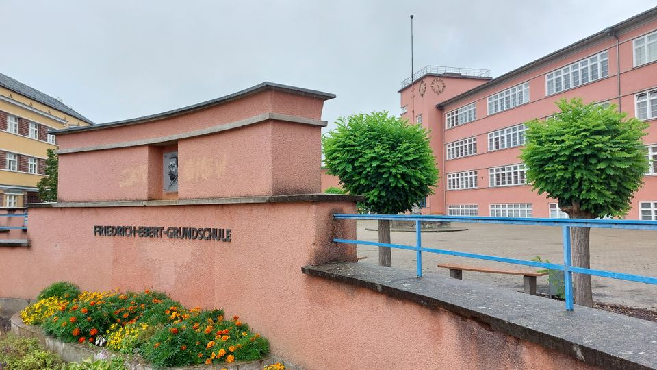 Außenansicht der Friedrich-Ebert-Grundschule mit einem rosa Gebäude im Hintergrund, zahlreichen rechteckigen Fenstern und einer Uhr an der Fassade. Im Vordergrund eine rosa Mauer mit einer Gedenktafel, darauf ein Blumenbeet und präzise geschnittene kleine Bäume.