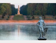 Steinfigur eines Mannes am Ufer eines Sees, im Hintergrund ein nebelbedeckter Park mit symmetrischen Wegen und einem Obelisken vor dichtem Wald.
