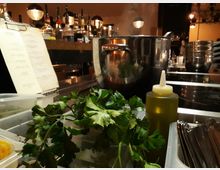 Eine Küchentheke mit Kochutensilien und Zutaten, darunter frische Petersilie, eine Flasche mit Öl, Löffel in Behältern und ein großer Edelstahltopf. Im Hintergrund stehen Flaschen und Speisekarten unter warmem Licht auf einer Bar.