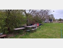 Eine Wiese mit mehreren Holztischen und -bänken nahe einer Hecke mit Bäumen. Im Hintergrund sind ein Schuppen, ein Trampolin und ein Zaun mit Pferdekoppel zu sehen.