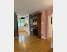 Wohnzimmer mit Holzboden, einem hölzernen Vitrinenschrank an einer rosa Wand und einer angrenzenden offenen Küche mit weißen Schränken und einem großen Fenster. Ein Weinregal steht an der grauen Wand neben der Küche.