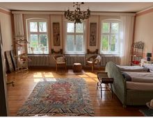 Ein gemütlich eingerichtetes Schlafzimmer mit Holzboden, einem großen bunten Teppich in der Mitte und einem Doppelbett mit einem grünlichen Korbgestell. Drei große Fenster lassen viel Tageslicht ein, dahinter ist eine grüne Landschaft sichtbar; daneben stehen Rattanstühle und Heizkörper unter den Fenstern. Im Raum befinden sich dekorative Elemente wie Bilderrahmen, Pflanzen, ein Kronleuchter und ein Kleiderständer.