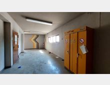 Ein leerer Raum mit grauen Wänden, einer Wandbemalung in Braun- und Gelbtönen, einem schmalen Fensterband auf der rechten Seite und einem orangefarbenen Sicherheitsschrank mit Warnzeichen. Eine geschlossene Holztür befindet sich auf der linken Seite, der Boden ist schlicht und abgenutzt.