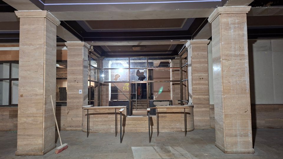 Innenansicht einer verlassenen Empfangs- oder Lobbyzone mit breiten steinverkleideten Säulen, dunkler Kassettendecke und einem gläsernen Durchgang in der Mitte. Vor dem Eingang stehen zwei schmale Tische; im Hintergrund sind beschädigte Fensterflächen, Staub und verstreute Gebäudeteile zu sehen.