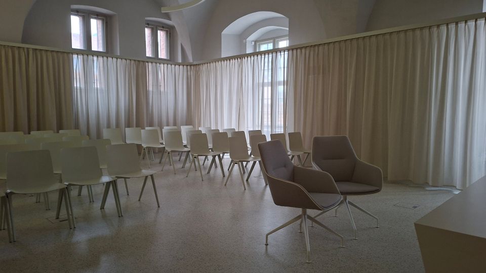Heller Veranstaltungs- oder Seminarraum mit hohen Gewölbedecken und mehreren Fenstern, umgeben von langen weißen Vorhängen als Raumtrennung. In der Mitte stehen Reihen weißer Stühle sowie vorne zwei graue Sessel auf hellem, glattem Boden.