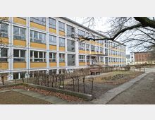 Mehrstöckiges Schul- oder Verwaltungsgebäude mit gelb-weißer Rasterfassade und vielen rechteckigen Fenstern, davor ein kleiner Hof mit Hecken, Wegen und einer Bank. Kahle Baumäste ragen über den Vorplatz, im Hintergrund sind weitere Gebäude zu sehen.