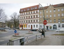 Eine Straßenszene mit mehrstöckigem Wohn- und Geschäftsgebäude im Stil eines Altbaus, mit weißer Fassade und roten Fensterrahmen. Im Vordergrund befinden sich Verkehrsinseln, ein "STOP"-Schild und Gehwege; die Straße trägt den Namen "Emmentaler Straße".
