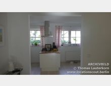 Moderne Küche mit einer freistehenden Kochinsel und hölzerner Arbeitsplatte, darüber eine Edelstahldunstabzugshaube. Im Hintergrund große Fenster mit rosa Vorhängen, Fensterbänke mit Pflanzen und Dekorationen.