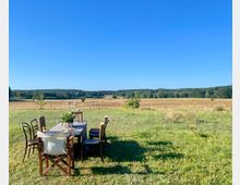 Ein einfacher Holztisch mit Stühlen steht auf einer Wiese, geschmückt mit Blumen und Trinkgläsern, vor einer weitläufigen Feldlandschaft mit Wäldern am Horizont unter klarem, blauem Himmel.