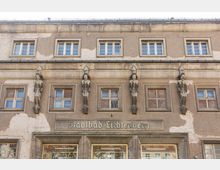 Fassade des Stadtbads Lichtenberg, einem historischen Gebäude mit Putzfassade, mehreren rechteckigen Fenstern und dekorativen Skulpturen in Frauenform, die als Stützen zwischen den Fenstern dienen. Unter den Fenstern der Erdgeschoss-Etage befindet sich der Schriftzug "Stadtbad Lichtenberg".